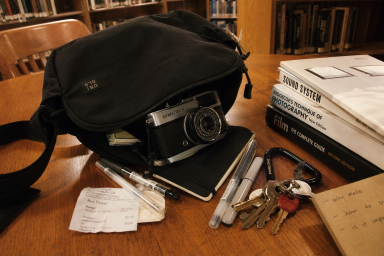A black camera bag with an Olympus trip camera on a table surrounded by pens, keys, a bus ticket, and photography books.