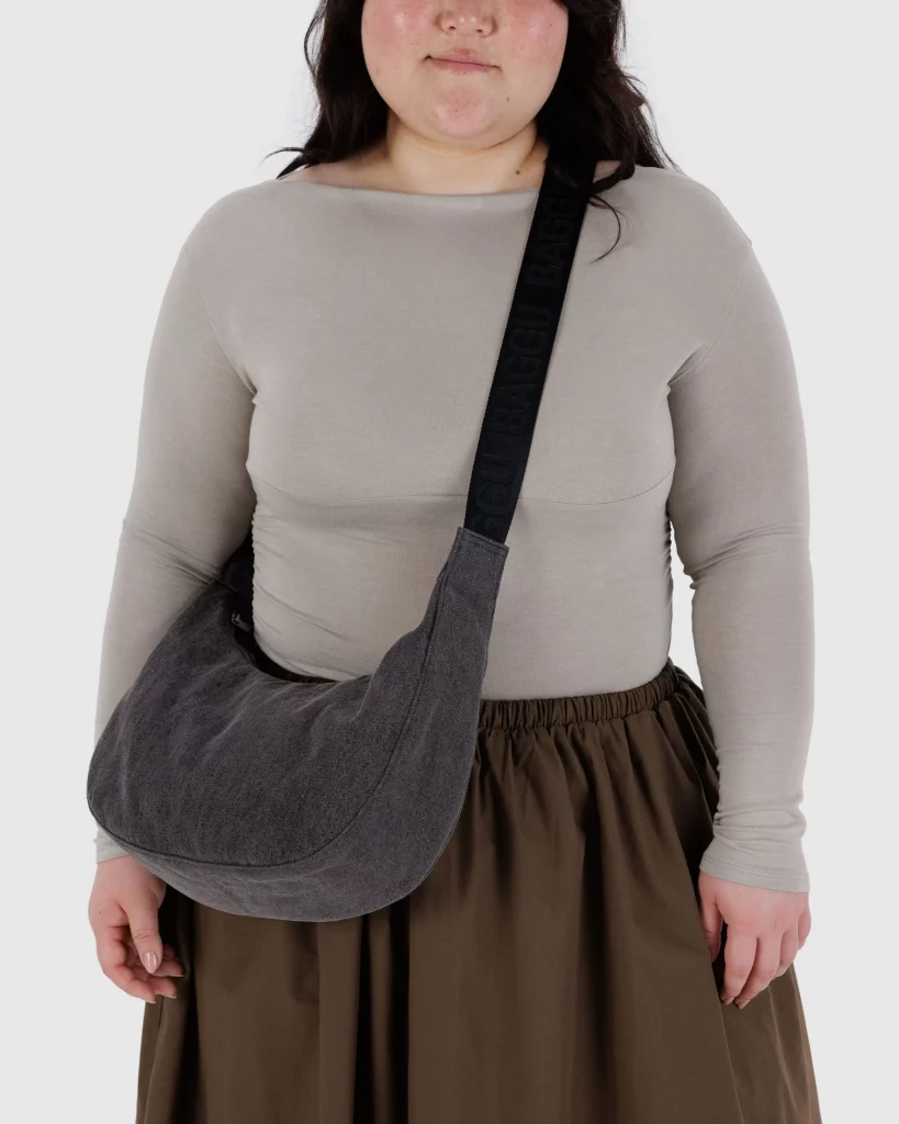 A woman wearing a beige long-sleeve top and a brown skirt, holding a dark grey bag.